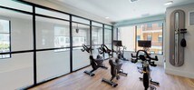 This well-appointed fitness center offers a comprehensive workout environment thoughtfully designed for a variety of training styles. The space is defined by warm hardwood-style flooring, exposed brick columns, and floor-to-ceiling glass partition walls framed in sleek black steel, lending an industrial-chic aesthetic throughout.

A dedicated cycling studio is enclosed by the glass partitions and outfitted with multiple stationary bikes, each equipped with individual monitors for on-demand class content. Generous windows along the exterior walls flood the studio with natural light and provide views of the surrounding building exterior.

The main workout area is open and spacious, featuring a well-stocked free weights section with a full rack of dumbbells, kettlebells, and medicine balls of varying sizes. A wall-mounted all-in-one training system adds versatile strength and conditioning options. Large mirrors expand the sense of space and support form-checking during workouts.

Near the entrance, a prominent digital display screen showcases available fitness app integrations, allowing users to track schedules, performance, and on-demand content. The transition areas between zones feature exposed brick accent columns that add texture and visual warmth. Recessed lighting throughout ensures consistent, even illumination across the entire facility. The overall design balances functionality and contemporary style, creating an inviting and motivating environment for daily fitness routines.