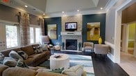 This thoughtfully designed living space showcases a harmonious blend of contemporary style and comfortable elegance. The room's most striking feature is the floor-to-ceiling mosaic tile surround framing a central fireplace, above which a flat-screen television is mounted, creating a natural focal point for the space. A tray ceiling adds architectural depth and dimension overhead, while recessed lighting provides a clean, modern illumination throughout.

The deep teal accent wall serves as a bold backdrop, complemented by warm earth-toned artwork that ties together the room's cohesive color palette of teals, golds, and rich browns. Dark hardwood flooring grounds the space and contrasts elegantly with the lighter furnishings.

Seating is arranged to encourage conversation, featuring a rich brown leather sofa adorned with decorative pillows, accent chairs in neutral toned upholstery, and additional seating near a window dressed with patterned drapes that allow natural light to filter in. A round upholstered ottoman serves as a central coffee table atop a watercolor-style area rug. Decorative vases and sculptural elements add curated finishing touches throughout.