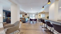 This single-floor space is thoughtfully designed with an open-concept layout that seamlessly connects multiple functional zones. The expansive main area flows between a casual lounge, a formal dining space, and a media room, all unified by warm hardwood-style flooring and crisp white walls.

The living area features a comfortable seating arrangement with a grey sectional sofa, coordinating armchairs, and a wooden coffee table, accented by colorful throw pillows and curated artwork on the walls. Adjacent to it, a dedicated media lounge boasts a bold teal accent wall, a large wall-mounted television, and a curved sectional sofa — ideal for relaxed gatherings.

The dining zone is anchored by a large rectangular table with ample seating, illuminated by a stylish multi-bulb chandelier. Natural light filters in through multiple windows and a sliding glass door that offers views of the surrounding outdoor environment. A high-top bar counter provides additional casual seating.

The fully equipped kitchen features white shaker cabinets, classic subway tile backsplash, dark countertops, and stainless steel appliances including a double oven, dishwasher, and a large French door refrigerator. Recessed and flush-mount ceiling lighting throughout the space ensures bright, consistent illumination in every area.