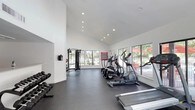 This single-floor fitness center offers a clean, modern environment designed to support a full range of workout routines. The space is characterized by soaring ceilings that lend an open, airy feel, while the crisp white walls and recessed lighting create a bright and energizing atmosphere throughout.

The main workout area is generously proportioned, providing ample room for multiple users to exercise simultaneously without feeling crowded. A row of dumbbell racks lines one wall, offering a variety of free weight options for strength training. A cable machine is positioned nearby, adding versatility for resistance-based exercises. The cardio section features multiple elliptical trainers and at least one treadmill, arranged to allow clear sightlines toward the outdoors.

Large floor-to-ceiling windows run along one side of the space, flooding the interior with natural light and offering views of an outdoor patio area with seating and lush surrounding greenery. This connection to the outdoors enhances the overall ambiance and creates an inviting setting for workouts. The dark rubberized flooring provides durability and traction while complementing the facility's contemporary aesthetic. An informational board is mounted on the wall near the cardio equipment, offering guidance for facility users. Overall, the space is thoughtfully designed for both function and comfort.