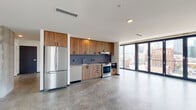 This contemporary urban unit is defined by its clean, modern aesthetic and an abundance of natural light streaming through expansive floor-to-ceiling windows and multi-panel glass doors that frame sweeping views of the city skyline. Polished concrete floors extend throughout the entire space, lending an industrial yet refined character that ties each area together cohesively.

The open-plan living and kitchen area is generously proportioned, with the kitchen positioned along one wall to maximize the sense of openness. The kitchen features warm wood cabinetry extending to the ceiling, complemented by a blue-grey mosaic tile backsplash, light-toned countertops, a stainless steel sink, and a full suite of stainless steel appliances including a refrigerator, dishwasher, and range with oven. Matte black hardware provides a sharp, modern accent throughout.

The living area benefits from multiple glass door panels that open to panoramic city views, with exposed concrete columns framing the glazing and reinforcing the building's structural character. A separate bedroom area also enjoys floor-to-ceiling corner windows offering additional skyline views.

The bathroom is boldly finished with a geometric patterned tile treatment in dark grey, white, and charcoal tones, a wall-mounted vanity with an open metal frame, a large mirror with strip lighting, and a glass-enclosed shower.