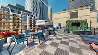 Located in Chicago's vibrant Streeterville neighborhood, this rooftop deck at 200 East Chestnut Street offers an expansive open-air retreat with sweeping views of the city skyline. The deck is finished with a distinctive checkered wood tile pattern that lends a stylish, contemporary feel to the outdoor space.

The rooftop is thoughtfully divided into multiple functional zones. A dedicated lounge area features modern outdoor sofas and chairs in teal and gray tones, anchored by a living green wall adorned with lush vertical foliage and a mounted flat-screen television — ideal for relaxed gatherings. Alongside this, a sunbathing zone is equipped with a row of cushioned chaise lounges, perfect for soaking in the ample natural sunlight.

The dining and entertaining section includes multiple round tables with teal sling chairs and vibrant potted floral arrangements that add pops of red and green throughout. Stainless steel grilling stations provide full outdoor cooking capabilities, making the space well-suited for social events and alfresco meals.

Perimeter railings frame the deck while keeping the dramatic panoramic views of Chicago's high-rises and architectural landmarks unobstructed on all sides. The combination of comfortable furnishings, greenery, entertainment amenities, and iconic urban vistas makes this rooftop an exceptional outdoor amenity space.