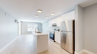 This well-designed 1-bedroom, 1-bathroom unit offers a thoughtfully laid-out single-floor living space with a clean, contemporary aesthetic throughout. The open-concept kitchen and living area serves as the heart of the home, featuring light hardwood-style flooring that extends seamlessly through the main living areas. The kitchen is outfitted with high-gloss white cabinetry, a large white quartz island with an undermount stainless steel sink, and a full suite of stainless steel appliances, including a refrigerator and electric range. Soft, neutral wall tones in muted grey and pale blue complement the bright, airy atmosphere. The living area benefits from natural light through a large sliding glass door leading to an outdoor space, as well as an additional window. The bedroom is a comfortable, well-proportioned room with the same light flooring and neutral tones, highlighted by a stylish black-hardware sliding barn door that opens to a dedicated walk-in closet with built-in shelving. The full bathroom features a tub-shower combination with a clean white surround, a white vanity, and neutral tile flooring. Flush-mount ceiling fixtures provide consistent, even lighting throughout the unit, reinforcing its bright and modern character.