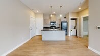Located at 2146 Heyboer Dr in Holland, Michigan, this single-floor unit offers a thoughtfully designed open-concept layout with a clean, contemporary aesthetic throughout. The kitchen serves as a central hub of the living space, featuring a large island with a double-basin sink and dark countertops, pendant lighting overhead, and white cabinetry extending to the upper walls. Black appliances — including a refrigerator, electric range, and over-the-range microwave — provide a bold contrast against the light cabinetry. Light wood-look flooring runs throughout the main living area, complementing the neutral beige and white tones of the walls and trim. The kitchen flows seamlessly into the living area, which is bathed in natural light from a set of glass French doors that open to the outdoors. A dedicated in-unit laundry area with a stacked washer and dryer adds everyday convenience. The bathroom is neatly finished with wood-look flooring, a vanity with cabinet storage, a lighted mirror, and clean white fixtures. The bedroom offers plush carpeting, warm neutral walls, and a broad window that invites ample natural light and views of the surrounding neighborhood, creating a calm and comfortable retreat.