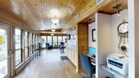 This single-floor space showcases a welcoming, lodge-inspired aesthetic defined by warm knotty pine wood paneling across both the walls and tongue-and-groove ceiling, complemented by light hardwood flooring throughout. Classic pendant and flush-mount ceiling lights provide a soft, inviting glow that pairs naturally with the ample daylight streaming in through a wall of large, multi-pane windows along the far end of the space.

The open layout flows seamlessly from a practical workstation area near the entrance — equipped with a desktop computer, granite-topped counter, and built-in cabinetry with light wood finishes — into a relaxed lounge and gathering area. The lounge is furnished with high-top bar tables and chairs alongside comfortable armchairs, making it suitable for both casual socializing and quiet individual use. A wall-mounted television adds a functional touch to the lounge zone.

Structural columns with white trim create subtle visual definition between areas without interrupting the open feel. The overall ambiance is warm, rustic yet polished, blending natural materials with practical amenities to create a comfortable and functional communal environment.