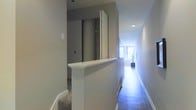 This single-floor space showcases a clean, contemporary design characterized by neutral grey walls, crisp white trim, and hardwood flooring that runs the length of a long, narrow hallway. Recessed ceiling lighting provides consistent illumination throughout the corridor, while a window at the far end draws in natural light, creating a bright and inviting focal point. A half-wall partition with a white-painted cap adds an architectural detail that subtly divides areas while maintaining an open visual connection between spaces. Adjacent to the partition, a sliding door in a darker grey tone offers access to a separate room or storage area, contributing a layered sense of depth to the layout. The transition between carpeted and hardwood flooring hints at distinct functional zones within the space. The overall ambiance is modern and understated, with a cohesive neutral palette that lends a calm, orderly atmosphere. The streamlined design details, including the clean baseboards, smooth wall finishes, and minimal ornamentation, reflect a thoughtfully executed interior that prioritizes both practicality and aesthetic simplicity.