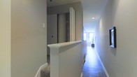 This single-floor space showcases a clean, contemporary design characterized by neutral grey walls, crisp white trim, and hardwood flooring that runs the length of a long, narrow hallway. Recessed ceiling lighting provides consistent illumination throughout the corridor, while a window at the far end draws in natural light, creating a bright and inviting focal point. A half-wall partition with a white-painted cap adds an architectural detail that subtly divides areas while maintaining an open visual connection between spaces. Adjacent to the partition, a sliding door in a darker grey tone offers access to a separate room or storage area, contributing a layered sense of depth to the layout. The transition between carpeted and hardwood flooring hints at distinct functional zones within the space. The overall ambiance is modern and understated, with a cohesive neutral palette that lends a calm, orderly atmosphere. The streamlined design details, including the clean baseboards, smooth wall finishes, and minimal ornamentation, reflect a thoughtfully executed interior that prioritizes both practicality and aesthetic simplicity.