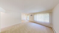 This 2-bedroom, 1-bathroom residence is thoughtfully organized across a single floor, making efficient use of its space. The open living room serves as the heart of the home, featuring wall-to-wall carpeting, baseboard heating, a ceiling fan, and a wall-mounted air conditioning unit. Large sliding glass doors lead to an outdoor balcony, while oversized vertical-blind-covered windows flood the room with abundant natural light and views of surrounding greenery.

The kitchen is compact yet functional, equipped with white cabinetry, a full-size refrigerator, an electric coil range with overhead ventilation, and ample counter space for everyday cooking needs.

Both bedrooms are carpeted and finished in neutral tones, creating a calm and restful atmosphere. Each room is fitted with a built-in closet featuring bifold doors with brass hardware, offering practical storage solutions. The primary bedroom is generously sized and receives natural light through a vertically-slatted window.

The single full bathroom includes a tiled tub-shower combination with classic square white tilework, chrome fixtures, and a clean, timeless aesthetic.

A hallway connects the private areas of the home, providing a clear and logical flow throughout. Baseboard heating is present throughout, ensuring consistent comfort year-round.