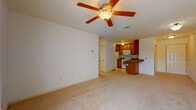 This single-floor unit offers a practical and comfortable layout designed for everyday living. The open-concept living area is finished with plush, neutral-toned carpet and features a ceiling fan with integrated lighting, creating a welcoming atmosphere. The space flows directly into the kitchen, which is defined by warm medium-toned wooden cabinetry, dark granite-style countertops, and a breakfast bar peninsula ideal for casual dining. White appliances — including a range, microwave, dishwasher, and refrigerator — complement the light tile flooring underfoot, and ample upper and lower cabinet storage ensures generous functionality. A separate bedroom, also carpeted, provides a quiet retreat with a closet and easy access back to the main living area. Sliding glass doors in the living space introduce natural light and provide access to an outdoor area, adding an airy quality to the interior. The bathroom is finished in crisp white, featuring a single-sink vanity with storage drawers, a large wall mirror framed by a Hollywood-style light bar, and an enclosed walk-in shower with glass sliding doors and tile flooring. Throughout the unit, neutral wall tones and consistent finishes create a clean, cohesive aesthetic.