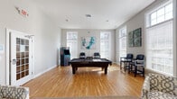 Located at 2067 Springdale Ln in Gallatin, Tennessee, this single-floor community amenity space is thoughtfully designed to offer residents a versatile environment for both recreation and leisure. The space is defined by an open, airy layout with neutral gray walls, warm hardwood flooring throughout the game area, and large windows dressed with white blinds that invite abundant natural light from multiple directions.

The centerpiece of the space is a full-size pool table positioned beneath a sleek pendant light fixture, complemented by a classic arcade cabinet that adds a fun, retro element to the room. High-backed spectator chairs are arranged near the windows, providing comfortable seating for onlookers. Abstract artwork adorns the walls, adding a touch of contemporary style throughout.

Transitioning into the lounge area, the flooring shifts to plush carpet, and the ambiance becomes noticeably more relaxed. A pair of patterned armchairs and a round side table create an intimate seating nook, while a teal accent wall adds a bold pop of color. A wall-mounted flat-screen television makes this corner well-suited for casual viewing or socializing. A glass-paneled door connects the areas and enhances the sense of openness. Fitness equipment is also present, rounding out this well-appointed community gathering space.