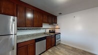 Located along VFW Parkway in the West Roxbury neighborhood of Boston, MA, this single-floor residence offers a well-appointed and functional layout. The kitchen is a standout feature, showcasing rich dark-wood cabinetry extending to the ceiling for ample storage, granite countertops, a classic white subway tile backsplash accented by a decorative mosaic border, and wood-look flooring. Stainless steel appliances — including an electric range and dishwasher — complete the space, making it both practical and visually appealing. The living and bedroom areas are finished with plush carpeting in a neutral blue-gray tone, lending a comfortable, quiet feel underfoot. Baseboard heating runs throughout, ensuring consistent warmth. The living area features a sliding glass door that opens to a private balcony with outdoor views, allowing natural light to fill the space. A wall-mounted air conditioning unit provides additional climate control. The bathroom retains a retro charm with a teal soaking tub, mosaic tile flooring, white subway wall tile with teal diamond accents, and a mirrored medicine cabinet above a clean vanity. Warm-toned wooden interior doors add a cohesive design element throughout the home.