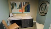 This single-floor space showcases a carefully considered interior design with a blend of modern aesthetics and functional elements. The space features warm, neutral wall tones complemented by teal-green accent walls, creating a layered and inviting color palette throughout. Durable wood-look flooring runs underfoot, providing a clean and cohesive foundation for the interior.

A standout feature is the bold, large-scale abstract artwork displayed prominently on the wall, incorporating vibrant hues of orange, green, blue, and cream — adding a dynamic visual focal point to the space. Below the artwork, a sleek built-in or freestanding cabinet unit with a textured dark wood-grain finish is topped with a light, veined stone-look surface, offering both storage and workspace functionality. The cabinetry features brushed metal hardware, contributing to the contemporary aesthetic.

Seating elements, including tan leather-toned stools or chairs, are positioned throughout, suggesting a space designed for comfort and interaction. Decorative circular mirrors further enhance the walls, adding depth and a sense of openness. Recessed electrical outlets are thoughtfully placed along the walls, ensuring practicality. Overall, the space presents a polished, professional environment with curated design details that balance form and function seamlessly.