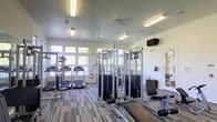 This single-floor fitness center offers a comprehensive workout environment thoughtfully designed to accommodate a range of exercise needs. The space is outfitted with multiple treadmills and cardio machines positioned along large, multi-pane windows that line the walls, flooding the interior with generous natural light and providing pleasant outdoor views while exercising. Ceiling fans are mounted overhead, contributing to air circulation and overall comfort throughout the workout area. A selection of cable-based strength training machines and resistance equipment occupies the central floor space, providing users with a variety of options for full-body conditioning. The flooring features a durable, patterned carpet in neutral tones, adding a finished look while providing underfoot comfort. Recessed and fluorescent lighting supplements the natural light for consistent visibility at all hours. A wall-mounted speaker is visible, suggesting the space is equipped for audio. A large mirror is positioned along one wall, a practical addition that supports proper form during exercise. The overall ambiance is clean, bright, and functional, making this fitness center a well-rounded facility suited for both casual and dedicated fitness enthusiasts.