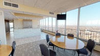 This elevated gathering space is designed to accommodate meetings, events, and social functions, offering an open, light-filled layout defined by expansive floor-to-ceiling windows that frame sweeping panoramic views of the surrounding cityscape and beyond. The generous main area is furnished with multiple round tables and padded chairs arranged throughout, providing flexible seating for groups of varying sizes. Patterned carpet covers much of the floor, while tiled sections near the service areas add contrast and practicality. A wall-mounted flat-screen television serves the seating area, making the space suitable for presentations or group viewings. One interior wall features a textured chevron-patterned finish adorned with a series of large-format cityscape artworks, adding a decorative focal point to the room. A dedicated kitchenette is equipped with warm wood cabinetry, granite countertops, a tile backsplash, stainless steel appliances including a microwave, dishwasher, and cooktop, along with a service counter finished in tile. Access to an outdoor terrace is available through the windowed perimeter, extending the usable space and offering open-air views. The drop ceiling with recessed lighting throughout keeps the ambiance bright and functional.
