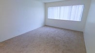 This single-floor space offers a clean, understated layout designed for comfort and functionality. The interior is defined by its neutral color palette, with crisp white walls that create a bright and airy atmosphere throughout. The main living area features wall-to-wall plush carpeting in a warm beige tone, providing a soft and inviting surface underfoot. A large window dressed with vertical blinds serves as a standout architectural feature, allowing generous amounts of natural light to fill the room while offering adjustable privacy. The smooth, light-colored ceiling complements the walls seamlessly, contributing to the open and uncluttered feel of the space. The overall ambiance is calm and versatile, providing a neutral canvas well-suited for a variety of interior design styles. The simplicity of the space lends itself to easy personalization, making it adaptable to a range of uses. The combination of ample natural light, soft flooring, and clean wall surfaces gives the interior a welcoming and practical character throughout.