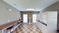 This single-floor shared laundry facility is designed with both functionality and comfort in mind. The room features a row of stacked, commercial-grade washer and dryer units along one wall, providing ample capacity for multiple users. A built-in countertop runs along the opposite wall, offering a generous folding and sorting surface complemented by bar-height seating — ideal for residents who wish to wait while their laundry cycles complete. A centrally positioned coin-operated payment kiosk provides convenient access to machine controls. The space is finished with a distinctive tan and white checkered tile floor, adding a classic, clean aesthetic to the utilitarian setting. Two windows fitted with blinds allow natural light to fill the room, creating a bright and airy atmosphere despite the functional nature of the space. The walls are painted in a neutral gray tone, and overhead fluorescent lighting ensures consistent illumination throughout. The overall layout is thoughtfully organized to maximize usability, making this laundry facility a practical and comfortable shared amenity.