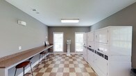 This single-floor shared laundry facility is designed with both functionality and comfort in mind. The room features a row of stacked, commercial-grade washer and dryer units along one wall, providing ample capacity for multiple users. A built-in countertop runs along the opposite wall, offering a generous folding and sorting surface complemented by bar-height seating — ideal for residents who wish to wait while their laundry cycles complete. A centrally positioned coin-operated payment kiosk provides convenient access to machine controls. The space is finished with a distinctive tan and white checkered tile floor, adding a classic, clean aesthetic to the utilitarian setting. Two windows fitted with blinds allow natural light to fill the room, creating a bright and airy atmosphere despite the functional nature of the space. The walls are painted in a neutral gray tone, and overhead fluorescent lighting ensures consistent illumination throughout. The overall layout is thoughtfully organized to maximize usability, making this laundry facility a practical and comfortable shared amenity.