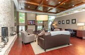Located at 42295 Parkside Cir in Sterling Heights, Michigan, this single-floor community space combines refined architectural design with a warm, welcoming ambiance. The main gathering area commands attention with its dramatic soaring ceilings adorned with rich, dark-stained coffered woodwork and recessed lighting, creating an atmosphere of elevated sophistication. A floor-to-ceiling stacked stone fireplace serves as the room's focal centerpiece, flanked by comfortable seating arrangements that include leather sofas, upholstered armchairs, and a central coffee table atop a woven area rug — ideal for relaxed socializing. Deep reddish-brown hardwood flooring flows throughout, complementing the warm, earthy color palette of taupe and grey walls. Large windows on multiple sides flood the interior with generous natural light while offering views of lush surrounding greenery. A built-in bookcase lines one wall, adding a library-like character to the lounge area, while a wall-mounted flat-screen television and a wet bar provide modern entertainment conveniences. A dedicated workstation area near the entrance features built-in desks with task lighting, catering to residents seeking a productive workspace. Framed artwork is thoughtfully displayed throughout, contributing to the space's curated, upscale aesthetic.
