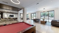 Located at 1110 Hammond Dr in Sandy Springs, GA, this single-floor community living space offers a generously scaled, open-plan layout designed for both relaxation and social engagement. The space is thoughtfully organized into several distinct zones that flow seamlessly into one another.

At the heart of the space is a game area anchored by a full-size billiards table, accented by suspended circular pendant lighting and a built-in banquette with upholstered seating along a textured feature wall. Adjacent to this is a lounge area with cream leather sofas and a dark marble coffee table, positioned beneath warm wood-panel tray ceilings with recessed lighting.

A dedicated media lounge features a wall-mounted television framed by a rich wood accent wall with decorative niches, flanked by plush neutral sofas and wood-plank flooring. A dining area with a natural wood table and upholstered chairs completes the interior entertaining zones.

Toward the exterior, a bright seating area with structured gray armchairs and a wood coffee table is bathed in natural light from expansive floor-to-ceiling black-framed glass doors and windows that open directly to an outdoor patio. A sculptural starburst chandelier adds a striking focal point overhead. The combination of large-format tile and warm hardwood flooring throughout ties the entire space together with a polished, contemporary aesthetic.