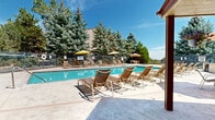 Located in Lakewood, Colorado, this outdoor recreational area offers a well-appointed pool and spa complex designed for relaxation and community enjoyment. The centerpiece is a generously sized, shallow swimming pool with crystal-clear turquoise water, complemented by a separate spa. The expansive concrete sundeck surrounds the pool on all sides, providing ample space for lounging in the abundant Colorado sunshine.

Numerous sling-style lounge chairs and seating arrangements are positioned throughout the deck, with several shaded options available beneath colorful market umbrellas and a covered pergola area adjacent to the clubhouse. An outdoor kitchen station equipped with a built-in BBQ grill offers convenient amenities for outdoor entertaining.

The pool area is fully enclosed by a sturdy black iron security fence, ensuring a safe and private environment. The surrounding landscape features mature evergreen and deciduous trees that provide natural shade and a scenic backdrop. The clubhouse building, finished in warm earth tones with red trim accents and large windows, anchors one side of the complex and provides access to restrooms and additional facilities. The overall setting evokes a resort-like atmosphere, ideal for enjoying the mild Colorado climate.