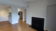 This single-floor space presents a straightforward and functional layout, beginning with an open living area that serves as the central hub of the home. A built-in fireplace with a clean white surround and dark firebox anchors one wall, adding a focal point and a source of warmth to the room. The living area is finished with warm hardwood-style flooring that extends throughout, contributing to a cohesive and inviting atmosphere. Neutral light gray walls keep the space feeling bright and versatile, complementing the overall clean aesthetic.

An open passageway connects the living area to the kitchen, which is visible from the main room. The kitchen features white cabinetry providing ample storage, and is equipped with a stainless steel refrigerator, offering a modern, functional cooking space. The flooring transitions seamlessly between the two areas, reinforcing the open feel of the layout. A ceiling vent is positioned to ensure climate control throughout. The combination of the fireplace, hardwood-style floors, and neutral palette gives the space a tidy, move-in ready character suited for everyday living.