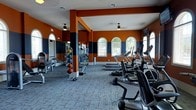 This fitness center makes a strong visual impression with its bold color scheme of deep orange and navy blue accent walls, creating an energetic atmosphere suited for an active workout environment. The single-story space features generous ceiling height, enhanced by ceiling fans that provide consistent airflow throughout the facility. Arched windows along one wall invite ample natural light while offering views of the surrounding outdoor area, contributing to an open and welcoming ambiance. The flooring consists of durable, textured carpet tiles designed to withstand heavy foot traffic. A mirrored wall on one side visually expands the space and allows users to monitor their form during exercise. The cardio area is well-stocked with a row of treadmills and elliptical machines positioned to take advantage of the natural light from the arched windows. The strength training section includes a medicine ball rack with a variety of weighted balls, weight benches, and additional resistance machines. Structural columns are integrated into the layout, subtly defining workout zones. Overall, the space is thoughtfully arranged to maximize functionality while maintaining an energetic and motivating environment.