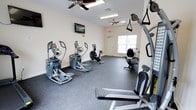 The fitness center at Windhurst Park Apartments is a well-equipped, single-floor exercise space designed to support residents' health and wellness routines. The room features durable rubber flooring suited for high-traffic use and is finished with clean white walls that contribute to a bright, open atmosphere. Two ceiling fans provide consistent air circulation throughout the space, keeping the environment comfortable during workouts.

The cardio section is prominently featured, housing multiple elliptical machines and a stationary air bike, offering a variety of low-impact aerobic options. A wall-mounted flat-screen television is positioned for easy viewing during cardio sessions. On the opposite side of the room, a cable resistance training machine with suspension straps and an adjustable workout bench provide options for strength and functional training exercises.

Natural light enters through a window near the far wall, adding to the room's welcoming feel. The layout is thoughtfully arranged to allow comfortable movement between equipment without feeling crowded. An emergency exit door is clearly marked, reflecting attention to safety standards. Overall, the fitness center offers a practical and accessible workout environment for residents.