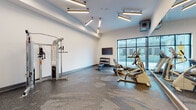 This contemporary fitness center in Baltimore, MD, is thoughtfully designed to support a wide range of workout routines across two connected zones. The main exercise area features durable speckled rubber flooring and crisp white walls, creating a clean, energizing environment. Distinctive linear pendant lighting is arranged in a starburst pattern across the ceiling, providing bright, even illumination throughout the space. Large floor-to-ceiling windows along one wall frame views of an outdoor courtyard, flooding the room with natural light and creating an open, airy atmosphere. The main area is equipped with a cable resistance machine, a multi-tier dumbbell rack with a weight bench, a treadmill, an elliptical trainer, a rowing machine, and a wall-mounted flat-screen television for entertainment during workouts. An adjoining studio room offers a dedicated space for functional training, featuring the same speckled rubber flooring, a full wall of mirrors that amplify the sense of space, a recumbent exercise bike, and multiple medicine ball racks stocked with a variety of weighted balls in assorted sizes. This secondary room also benefits from its own climate control and direct access to the main gym area through a connecting door, making it a versatile space for stretching, balance training, or low-impact exercise.