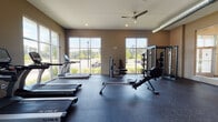 Located at 5801 Zebulon Rd in Macon, Georgia, this single-floor fitness center is designed to provide a well-equipped and comfortable workout environment. The space is divided into distinct exercise zones, each thoughtfully appointed with professional-grade equipment. The cardio area features a lineup of TRUE treadmills, ellipticals, and stationary bikes positioned alongside expansive floor-to-ceiling windows that flood the room with natural light and offer views of the surrounding landscape. A Bodycraft rowing machine and additional cable and weight machines round out the strength training offerings, while a dedicated multi-functional cable station and adjustable weight bench provide versatility for a full-body workout. Dumbbell racks and a free-standing cable crossover machine add further options for resistance training. The flooring transitions between durable rubber matting in the heavy equipment zone and carpet tile in the stretching and open exercise areas, providing comfort and safety throughout. Exposed ductwork and ceiling fans contribute to an industrial-modern aesthetic, while recessed lighting and a wall-mounted television enhance the functionality of the space. A built-in water fountain station ensures convenient hydration, and glass-panel entry doors connect the fitness center to adjacent amenity areas. Large windows throughout maintain a bright, energizing atmosphere ideal for an active workout experience.