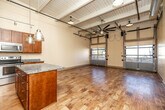 This single-floor space showcases an industrial-chic aesthetic defined by soaring exposed beam ceilings with white-painted wood planking, track lighting on overhead rails, and oversized pendant lights throughout. The open main living area is expansive and flexible, anchored by warm hardwood flooring that runs throughout. A standout feature is the full-width glass-panel roll-up garage door that floods the space with natural light and provides direct outdoor access, enhancing the loft's airy, versatile character. A large ceiling fan with additional pendant lighting completes the industrial look overhead.

The kitchen is well-appointed with rich cherry wood cabinetry extending to the ceiling, granite countertops, a central island with matching granite, stainless steel appliances including a refrigerator, range, microwave, and dishwasher, plus track lighting above. The layout offers generous prep and storage space.

A utility area adjacent to the bathroom houses a stacked washer and dryer for added convenience. The bathroom features light ceramic tile flooring, tile-surround tub and shower combo, a toilet, and a wood vanity. The overall ambiance is open, modern, and highly functional, suited for a variety of uses.