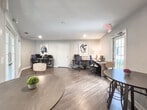 Located at 2255 Satellite Blvd in Duluth, Georgia, this single-floor business center offers a well-appointed and functional workspace designed for productivity and convenience. The room features warm hardwood-style flooring throughout, complemented by neutral gray walls and clean white trim that create a bright, professional atmosphere.

The workspace is anchored by a curved granite-top desk positioned along one wall, accommodating two computer workstations complete with ergonomic mesh task chairs and a printer, making it well-suited for residents or guests needing to handle business tasks. Framed abstract artwork adorns the walls, lending a polished, finished feel to the interior.

A casual high-top table with industrial-style swivel stools provides an additional spot for informal meetings or collaborative work. Natural light enters the space through multiple windows, including a full-lite glass exterior door that opens directly to the outdoors, offering views of the surrounding greenery and parking area.

A set of elegant French doors with gridded glass panels connects the business center to an adjacent common area, enhancing the sense of openness and allowing borrowed light from neighboring spaces. Recessed lighting and a ceiling vent ensure the room remains well-lit and comfortably climate-controlled. The overall ambiance is clean, modern, and inviting.