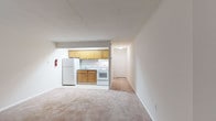 This single-floor unit offers a practical and functional layout centered around an open-concept living and kitchen area. The spacious main living area is finished with wall-to-wall beige carpeting and neutral white walls, creating a clean and versatile backdrop for furnishings. A wall-mounted air conditioning unit and a baseboard heater provide year-round climate control, while a private entry door with a window allows natural light into the space.

The kitchen is seamlessly integrated into the living area via a half-wall partition, providing an open feel while maintaining a defined cooking space. It features warm honey-toned oak cabinetry both above and below the counter, a light-toned laminate countertop with a single-basin stainless steel sink, a white electric coil-burner range with an overhead exhaust hood, and a full-size white refrigerator. The kitchen floor is finished in light-toned vinyl for easy maintenance.

The bathroom is finished in a crisp, all-white palette, featuring a full-size soaking tub with white square ceramic tile surround extending to the ceiling, a toilet, and an over-toilet storage cabinet. The clean, minimal design gives the bathroom a bright and tidy appearance. Overall, the unit presents a straightforward, well-maintained living space with all essential amenities included.