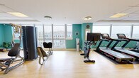 This fitness center offers a spacious, open-plan layout designed to accommodate a full range of workout activities. The space is defined by its vibrant teal accent walls, crisp white trim, and light hardwood flooring, creating an energetic yet clean aesthetic throughout.

Large floor-to-ceiling windows line one side of the room, flooding the interior with abundant natural light and offering expansive views of the surrounding urban cityscape. A full-length mirror wall further enhances the sense of openness and allows users to monitor their form during exercise.

The facility is well-appointed with a comprehensive selection of equipment, including treadmills, stationary spin bikes, a multi-station cable and strength machine, rowing and weight bench stations, and free weights organized neatly on a dedicated rack. Stability balls, medicine balls, and resistance tools are also available, supporting functional and flexibility training.

Recessed and flush-mounted ceiling lighting supplements the natural light, ensuring the space remains bright and well-illuminated throughout the day. A water dispenser station is conveniently positioned within the room. The facility is accessed through double wood-and-glass doors, and the open floor plan allows for comfortable circulation between equipment zones, making this fitness center a practical and inviting space for a wide variety of training needs.