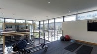 This contemporary fitness space is thoughtfully designed to create an energizing and motivating workout environment. The facility is defined by its expansive floor-to-ceiling windows that wrap around the perimeter, flooding the interior with natural light and offering unobstructed views of the surrounding greenery and outdoor terrace. The open-plan layout provides generous room for a wide variety of exercise equipment, including rowing machines, benches, and cardio stations, all arranged to maximize usability and flow. A dedicated stretching and mat area is equipped with stability balls and balance equipment, catering to flexibility and functional training routines. Sleek glass railings and pendant lighting add refined architectural detail, while recessed ceiling fixtures ensure consistent illumination throughout. The dark-toned carpet flooring offers both comfort and durability underfoot, complementing the modern, neutral color palette of the space. Large mirrors further enhance the sense of openness while serving a practical function for form checking during workouts. Overall, the facility strikes a balance between functional performance and sophisticated design, creating a welcoming and well-equipped environment for a full range of fitness activities.