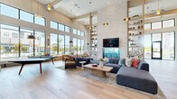 Located at 1830 Homerun Rd in Kissimmee, Florida, this single-story community clubhouse offers a generously sized, open-plan interior designed with a modern coastal aesthetic. The space is defined by dramatically soaring ceilings that amplify the sense of openness throughout, while light-toned hardwood flooring runs seamlessly across the entire floor plan, tying each area together with warmth and continuity.

At the heart of the space, a striking floor-to-ceiling white stacked-stone accent wall frames a built-in media display, flanked by open shelving adorned with curated décor and greenery. A large sectional sofa anchors the central lounge area, complemented by woven accent chairs and a sculptural coffee table atop a layered area rug.

An integrated wet bar with a blue glazed tile backsplash, under-counter refrigeration, draft taps, and a vertical wine rack adds a social and functional focal point. A nearby dining area with a bold table and colorful seating serves as a casual gathering spot.

Expansive floor-to-ceiling windows and glass doors flood the space with abundant natural light and offer unobstructed views of the outdoor pool area, lush landscaping, and swaying palms. A built-in window bench with colorful accent pillows provides additional relaxed seating alongside the sun-drenched perimeter. The overall ambiance is bright, airy, and inviting.