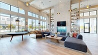 Located at 1830 Homerun Rd in Kissimmee, Florida, this single-story community clubhouse offers a generously sized, open-plan interior designed with a modern coastal aesthetic. The space is defined by dramatically soaring ceilings that amplify the sense of openness throughout, while light-toned hardwood flooring runs seamlessly across the entire floor plan, tying each area together with warmth and continuity.

At the heart of the space, a striking floor-to-ceiling white stacked-stone accent wall frames a built-in media display, flanked by open shelving adorned with curated décor and greenery. A large sectional sofa anchors the central lounge area, complemented by woven accent chairs and a sculptural coffee table atop a layered area rug.

An integrated wet bar with a blue glazed tile backsplash, under-counter refrigeration, draft taps, and a vertical wine rack adds a social and functional focal point. A nearby dining area with a bold table and colorful seating serves as a casual gathering spot.

Expansive floor-to-ceiling windows and glass doors flood the space with abundant natural light and offer unobstructed views of the outdoor pool area, lush landscaping, and swaying palms. A built-in window bench with colorful accent pillows provides additional relaxed seating alongside the sun-drenched perimeter. The overall ambiance is bright, airy, and inviting.