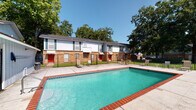 This outdoor pool facility in Houma, Louisiana, offers a generously sized swimming pool as its central feature. The pool is finished with classic brick coping along its perimeter and filled with clear turquoise water, providing a welcoming recreational environment. A concrete pool deck surrounds the water, furnished with seating areas that allow residents to relax under open skies framed by mature trees.

The entire pool area is enclosed by a chain-link fence, providing a secure boundary while maintaining visibility of the surrounding grounds. Entry points are integrated into the fence line, with metal handrails installed at the pool steps for safe access. Lush green grass borders the deck, and the backdrop features a two-story brick residential building with white siding accents, teal shutters, and distinctive red entry doors, contributing to a cohesive community setting.

Adjacent to the pool is a covered single-story utility structure with light gray vertical board siding, a coffered ceiling, and overhead lighting. This space houses a beverage vending machine and includes a small interior room accessible via a standard door, with a pass-through window. The concrete floor of this covered area serves as a practical transitional space connecting the pool zone to the broader property grounds.