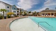 Located at 50 Newport Blvd in Port Wentworth, Georgia, this outdoor amenity space showcases a generously sized resort-style swimming pool as its centerpiece. The pool features a curved, freeform design with a shallow entry area, making it accessible and inviting for a wide range of guests. The surrounding deck is finished with attractive brick paver detailing, providing a durable and visually appealing surface throughout the space.

An abundance of white lounge chairs lines the pool perimeter, offering ample seating for sunbathing and relaxation. Shaded seating areas with umbrellas provide respite from the Georgia sun, while a dedicated pool house structure offers convenient access to facilities and posted pool guidelines. The entire pool area is enclosed by a black iron fence, ensuring privacy and security.

The landscaping features a mix of mature palm trees and tall pines, lending a coastal and resort-like ambiance to the setting. Open views of the surrounding greenery and wide Georgia skies enhance the sense of openness. The multi-story residential buildings adjacent to the pool are clad in classic coastal-inspired siding in soft blues and grays, complementing the overall aesthetic of the community and creating a cohesive, well-designed outdoor retreat.