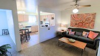 This single-story space offers an efficient and practical layout designed to maximize functionality. The living area features neutral-toned carpeted flooring and is anchored by a ceiling fan overhead. A wood-framed coffee table and dark upholstered sofa create a comfortable seating arrangement, while decorative wall art adds a touch of color to the space. A pass-through window connects the living area to the kitchen, creating an open and communicative flow between the two spaces.

The kitchen is outfitted with light wood cabinetry, a white refrigerator, and a range, providing essential appliances for everyday use. A window above the sink allows natural light to brighten the workspace. Adjacent to the kitchen, a dining nook accommodates a compact table with bar-style seating, making it well-suited for casual meals.

The overall ambiance is straightforward and functional, with light-colored walls throughout that contribute to a bright and airy feel. The layout is well-organized, ensuring easy movement between the living, dining, and kitchen areas, making the space practical for everyday living.
