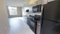 This Type 'B' studio unit offers a well-designed single-floor layout that efficiently combines living and kitchen functions in one open space. The kitchen is positioned along one wall and is outfitted with a full suite of black appliances, including a refrigerator, electric range, built-in microwave, and dishwasher. Dark granite countertops provide a sleek contrast against the contemporary dark-toned cabinetry, which features brushed metal hardware and extends to the upper wall for generous storage capacity. A single-basin sink with a modern faucet completes the kitchen area.

The open-concept living area flows directly from the kitchen, providing a sense of spaciousness despite the compact footprint. Light wood-toned flooring runs throughout, contributing to a clean, modern aesthetic. A large window along the far wall admits abundant natural light, brightening the neutral gray walls and enhancing the overall airiness of the space. The unit includes one full bathroom. The straightforward, functional layout makes efficient use of every area, offering a practical and modern living environment.