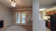 This single-story residence showcases a practical and open interior layout designed for comfortable everyday living. The main living area is anchored by a set of French doors that fill the space with natural light while providing a direct view of the outdoors. Horizontal blinds on the doors and the adjacent window offer privacy and light control as needed. The living area is finished with wall-to-wall carpeting in a neutral tone, complementing the warm beige walls throughout.

A ceiling fan with a built-in light fixture is centrally positioned in the living room, providing both air circulation and ambient lighting. Architectural interest is added by a structural column and a decorative beam detail near the ceiling, which subtly defines the transition between spaces.

A notable feature is the pass-through window cutout in the partial wall separating the living area from the kitchen, enhancing the open feel and allowing easy communication between rooms. The kitchen area, partially visible through the pass-through, is illuminated by a flush-mount ceiling light and includes a countertop ledge that doubles as a convenient serving or casual dining surface. Overall, the space presents a clean, functional layout with cohesive neutral finishes throughout.
