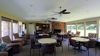 This single-story community room is thoughtfully designed to accommodate a variety of social gatherings and events. The open-plan layout is anchored by a striking stone fireplace that serves as a dramatic focal point, adding warmth and character to the overall ambiance. Multiple round tables surrounded by cushioned chairs are arranged throughout the room, providing ample seating for groups of varying sizes.

Overhead, several ceiling fans ensure comfortable air circulation throughout the space. Recessed lighting complements the abundant natural light that streams in through large sliding glass doors and windows lining one wall, offering pleasant views of the surrounding greenery outdoors.

At the back of the room, a kitchenette area features dark wood cabinetry, a refrigerator, and counter space — well-suited for catering or refreshment service during events. Additional lounge-style seating with upholstered armchairs is positioned throughout the room, encouraging informal conversation and relaxation.

The neutral color palette of beige walls and gray carpeted flooring creates a calm, inviting atmosphere. A dedicated door along the side wall provides convenient access to an adjacent area or restroom. Overall, this space balances functionality with a warm, communal aesthetic, making it an ideal venue for social events, meetings, or casual gatherings.