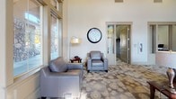 This single-floor amenity space is thoughtfully designed to serve as a vibrant social hub, combining comfort, functionality, and entertainment in one open layout. The grand lounge area anchors the space with soaring ceilings, an abundance of natural light streaming through large floor-to-ceiling windows, and plush gray leather seating arranged on patterned carpeting. Transom windows and interior glass panels add architectural character while maintaining visual openness throughout.

The fully equipped kitchen features rich dark cabinetry, granite countertops, a mosaic tile backsplash, stainless steel appliances including a side-by-side refrigerator, and wood-look flooring—making it well-suited for catered events or community gatherings. A checkered accent wall adds a modern design touch to this functional area.

Adjacent to the lounge, a dedicated game room provides entertainment options including a foosball table and a full-size billiards table, complemented by decorative poker and billiards-themed wall art. A high-top dining table positioned near a window overlooking the landscaped exterior grounds adds a casual seating option. Throughout the space, decorative mirrors, framed artwork, and warm accent lighting contribute to a welcoming and well-appointed ambiance suited for both relaxed gatherings and organized community events.