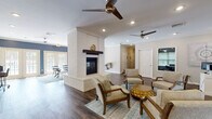 This single-story community clubhouse is designed to serve as a welcoming, multi-functional gathering space. The centerpiece of the main area is a striking white-painted brick fireplace with a rustic wood mantel, which anchors the open-plan layout and provides a natural division between two distinct lounge zones. One side features a seating arrangement with upholstered arm chairs arranged around a gold accent table atop a blue patterned area rug, while the other accommodates a glass-top dining table with mid-century modern chairs. Rich, dark hardwood-style flooring runs throughout, complemented by recessed lighting and multiple ceiling fans that promote comfort and airflow. French doors along one wall allow natural light to fill the space and provide views of an outdoor area. A workstation area with a desk and computer offers a practical resource for residents. The kitchen is outfitted with white cabinetry, a full-size refrigerator, microwave, and a peninsula counter with bar seating, ideal for casual gatherings. Adjacent to the main lounge, the fitness center is well-equipped with cardio machines, a cable weight system, a dumbbell rack, stability balls, wall-mounted mirrors, and mounted televisions, creating a comprehensive exercise environment within the facility.