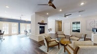 This single-story community clubhouse is designed to serve as a welcoming, multi-functional gathering space. The centerpiece of the main area is a striking white-painted brick fireplace with a rustic wood mantel, which anchors the open-plan layout and provides a natural division between two distinct lounge zones. One side features a seating arrangement with upholstered arm chairs arranged around a gold accent table atop a blue patterned area rug, while the other accommodates a glass-top dining table with mid-century modern chairs. Rich, dark hardwood-style flooring runs throughout, complemented by recessed lighting and multiple ceiling fans that promote comfort and airflow. French doors along one wall allow natural light to fill the space and provide views of an outdoor area. A workstation area with a desk and computer offers a practical resource for residents. The kitchen is outfitted with white cabinetry, a full-size refrigerator, microwave, and a peninsula counter with bar seating, ideal for casual gatherings. Adjacent to the main lounge, the fitness center is well-equipped with cardio machines, a cable weight system, a dumbbell rack, stability balls, wall-mounted mirrors, and mounted televisions, creating a comprehensive exercise environment within the facility.