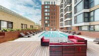 Located at 8021 Georgia Ave in Silver Spring, Maryland, this outdoor amenity space is a well-appointed courtyard retreat nestled within a contemporary multi-story residential building. The architecture features a combination of classic red brick and modern smooth façades, with large grid-style windows that frame the courtyard from multiple levels above.

At the heart of the space is a generously sized rectangular swimming pool with clear blue water, framed by a broad pool deck with rows of sleek chaise lounges positioned for sunbathing. The surrounding deck is finished in warm-toned hardwood planking, lending a refined, resort-like character to the outdoor environment.

Multiple seating areas are distributed throughout the deck, including wicker sectional sofas with bold red cushions and additional lounge chairs with neutral upholstery, providing varied options for relaxation and social gathering. Raised brick planter beds line portions of the perimeter, adding greenery and defining the space's boundaries with architectural consistency.

The courtyard benefits from open sky exposure, allowing ample natural light throughout the day. The surrounding building rises several stories, offering a sense of enclosure while maintaining an open, airy atmosphere. The overall design balances urban density with comfortable outdoor living, making this a highly functional and visually appealing amenity space in the heart of Silver Spring.
