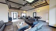 This single-floor communal space is designed to accommodate a variety of activities within a well-organized, open layout. The room's most notable architectural feature is its elevated tray ceiling, accented with dark trim detailing that adds a sense of depth and sophistication to the interior. Recessed lighting throughout the ceiling is complemented by a hanging drum pendant light, ensuring the space is well-illuminated.

The lounge is divided into distinct functional zones. A central seating area anchors the room, featuring dark leather sofas arranged around a low rectangular coffee table atop a patterned area rug, creating an inviting space for relaxation or casual conversation. Adjacent to this is a high-top table with bar-height seating, ideal for informal dining or group gatherings.

Toward the entrance, a dedicated workstation area includes desks equipped with computers, providing a practical space for productivity. The glass-paneled entry doors allow natural light to filter in, enhancing the bright, neutral color palette of the walls. Teal accent details on the doors and baseboards add a cohesive design touch throughout the space.