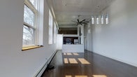 This single-floor space offers an open, airy layout defined by clean white walls and rich dark hardwood floors that stretch throughout the main living area. Soaring ceilings lend an impressive sense of volume, enhanced by a large ceiling fan that anchors the space. A row of tall windows lines one wall, flooding the interior with generous natural light and casting warm patterns across the floor.

At the heart of the space, a half-wall partition defines the kitchen area without sacrificing the open feel. The kitchen is equipped with stainless steel appliances, including a range and microwave, and features dark cabinetry that provides a striking contrast to the surrounding white walls. A cluster of elegant pendant lights with glass fixtures hangs above, adding a refined decorative touch.

Baseboard heating runs along the windowed wall, ensuring year-round comfort. The open-plan design seamlessly connects the living and kitchen areas, making the space highly functional and versatile. The combination of industrial-inspired elements — such as the exposed ceiling details — with warm hardwood flooring and abundant natural light creates a balanced, modern ambiance that is both practical and visually inviting.