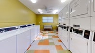 This shared laundry facility in Phenix City, Alabama is designed for practicality and comfort. The single-floor space is characterized by vibrant yellow walls and a colorful checkerboard tile floor in orange, green, and white, creating a lively and welcoming atmosphere. Bright overhead fluorescent lighting, supplemented by a ceiling fan, ensures the room is well-lit and comfortable throughout the day. Natural light also filters in through multiple windows fitted with white horizontal blinds, adding to the airy feel of the space.

The facility is generously outfitted with a row of commercial-grade top-load washing machines along one wall, and a bank of stacked commercial dryers along the opposite side, providing ample capacity for multiple users simultaneously. A long white folding table runs along one wall, offering convenient space for sorting and folding laundry. A classic white and wrought iron bench is positioned centrally, providing a comfortable waiting area. An additional folding or utility table near the entrance adds further functionality. Safety is addressed with a prominently mounted fire extinguisher near the exit door, which features a sidelight window for added visibility. The layout is open and efficient, making this laundry facility a highly functional communal amenity.