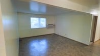 This single-floor space presents a straightforward, open layout with a clean and functional design. The main room is generously sized, offering an open feel accentuated by smooth white walls and a flat ceiling with a subtle recessed upper section that adds a modest architectural detail.

The flooring throughout consists of durable tile in a neutral gray tone, providing a practical and easy-to-maintain surface. Natural light enters through a single window fitted with vertical blinds and a security bar, illuminating the room adequately while maintaining privacy and security.

Climate control is handled by a wall-mounted through-the-wall air conditioning unit positioned directly beneath the window, offering both heating and cooling functionality. The baseboards are clean and consistent throughout, contributing to a tidy overall appearance.

The space is currently vacant, making it straightforward to assess its dimensions and versatility. The neutral color palette and open floor plan provide flexibility for a variety of uses or furniture configurations. Electrical outlets are visible along the walls, ensuring practical utility throughout the space.